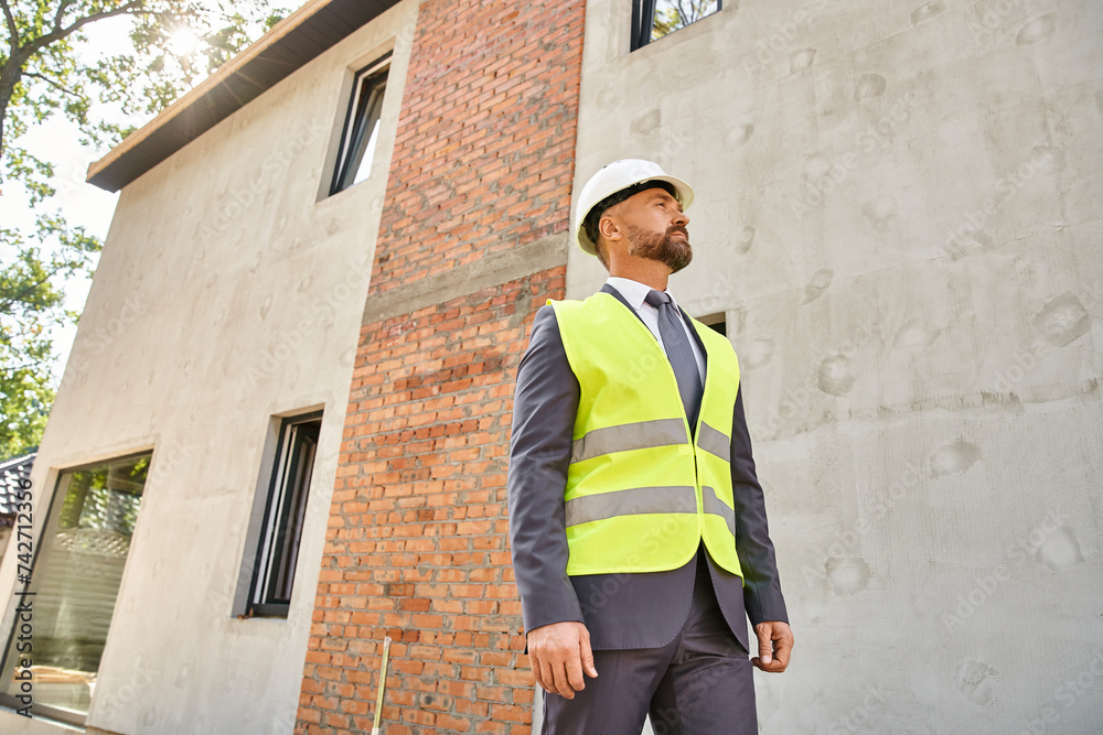 devoted businessman in safety vest and helmet posing on construction site and looking away devoted businessman in safety vest and helmet posing on construction site and looking away