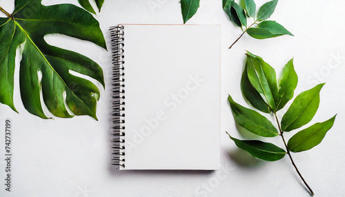Empty rustic style notepad with green leaves on a table. Blank white mockup sketchbook on white background, free space. Top view, copy space for text or advertisement.