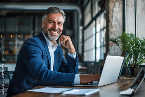 Happy smiling middle aged professional business man company executive ceo manager wearing blue suit sitting at desk in office working on laptop computer laughing at workplace, Generative AI