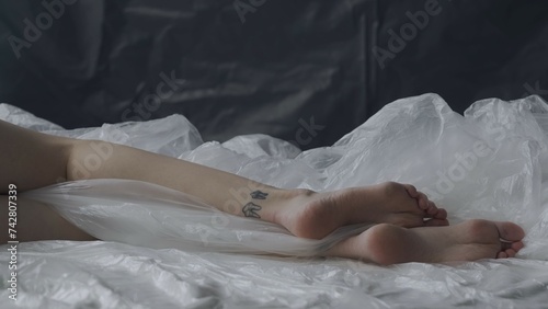 Female laying on the bed in the studio. Young woman legs feet close up seminude laying under thin sheet of oilcloth wrap.