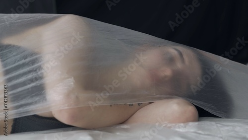 Female in underwear laying on the bed in the studio. Young woman seminude in lingerie laying posing under thin sheet of oilcloth wrap.
