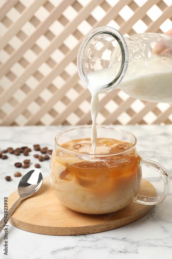 Woman pouring milk into glass with refreshing iced coffee at white marble table, closeup
