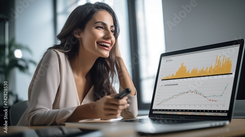 Smiling CEO brunette senior exectuive business woman in light office at her desk and laptop with growth graphs as financial stock or banking manager at work photo