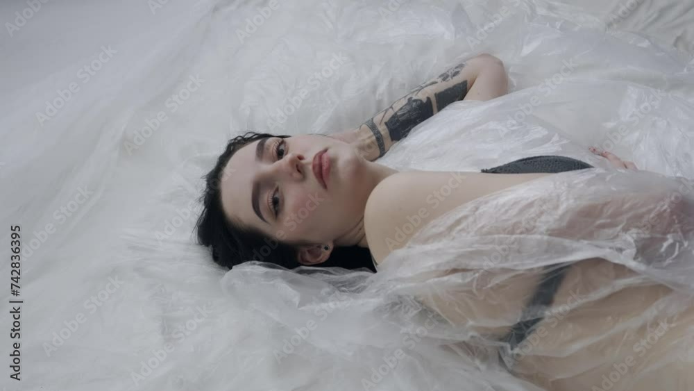 Female in underwear laying on the bed in the studio. Young woman seminude in lingerie laying posing under thin sheet of oilcloth wrap, looking at camera. Female in underwear laying on the bed in the studio. Young woman seminude in lingerie laying posing under thin sheet of oilcloth wrap, looking at camera.