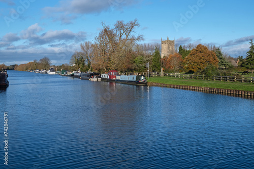 Scene on a sunny Autumn day with barges and houseboats moored on the Gloucester and Sharpness canal at Frampton on Severn UK. The 14th century parish church of St Mary sits on the bank of the canal