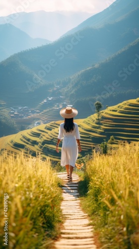Rear view of a Vietnamese girl in a long white dress and hat walking on a mountain and rice terraces on a farm.