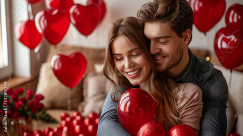 Happy Valentine's Day. Young couple in love holding a heart-shaped balloon, hiding behind it while sitting on the sofa in the living room at home. Romantic evening together.