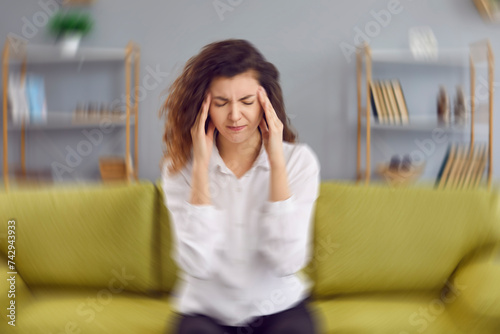 Woman feels dizzy and sits down on the couch. Young woman sitting on the sofa at home, feeling pain and a spinning sensation in her head. Concept of headache, vertigo, health problem, brain tumor photo