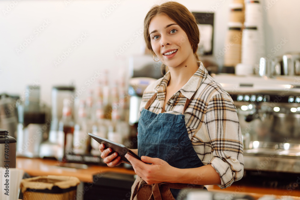 Smiling female barista takes an order from a tablet while standing at the bar counter in a coffee shop. Online order. Small business, people, takeaway and service concept. Smiling female barista takes an order from a tablet while standing at the bar counter in a coffee shop. Online order. Small business, people, takeaway and service concept.