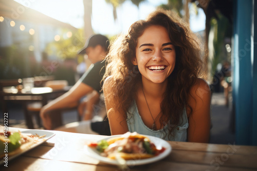 American young woman smiling while sitting outside at a table on a restaurant patio and eating delicious tacos.