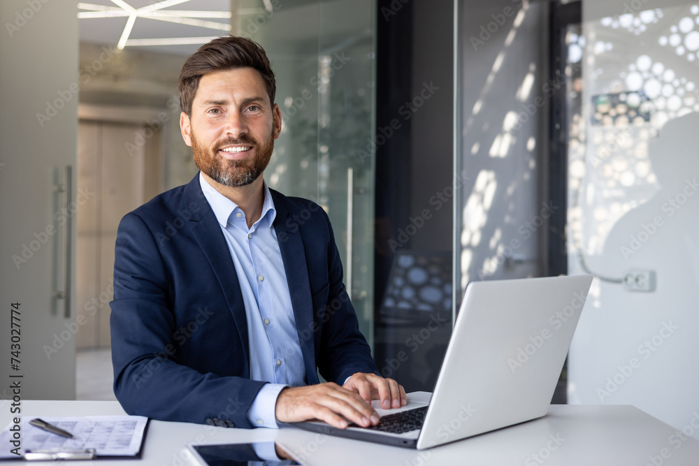 Portrait of a successful young businessman in a suit sitting at a desk in the office, working with documents and using a laptop, confidently and smilingly looking at the camera Portrait of a successful young businessman in a suit sitting at a desk in the office, working with documents and using a laptop, confidently and smilingly looking at the camera