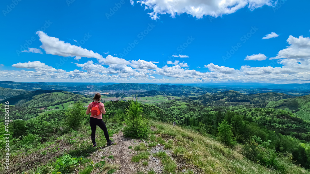 Hiker woman on rock formation along idyllic hiking trail through lush green forest in Grazer Bergland, Prealps East of the Mur, Styria, Austria. Vibrant green leaves. Soft hills in alpine landscape