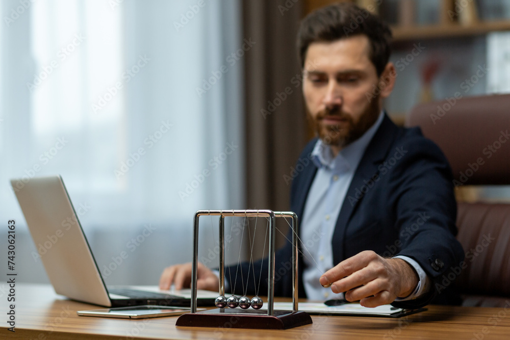 A serious and successful young man sitting in the office at a desk in a suit uses Newton's Cradle by launching balls with his hand A serious and successful young man sitting in the office at a desk in a suit uses Newton's Cradle by launching balls with his hand