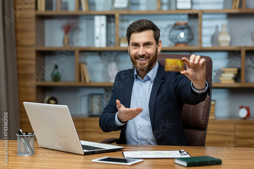 Portrait of a smiling young successful businessman, banker, financial director sitting at an office desk in a suit, holding and showing a golden plastic credit card to the camera photo