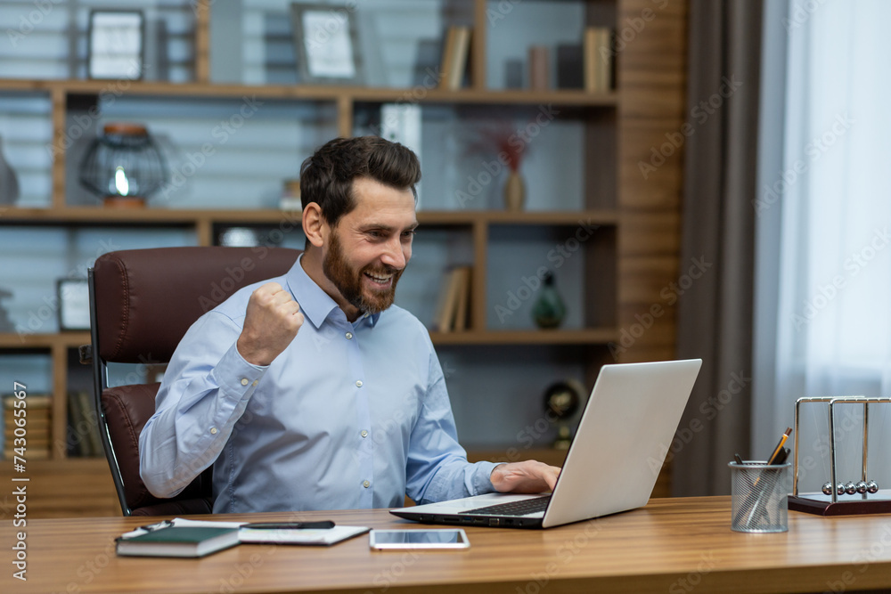 Happy young business man sitting in the office at the desk with a notebook and rejoices at the success, looking at the window, showing a victory gesture with his hand