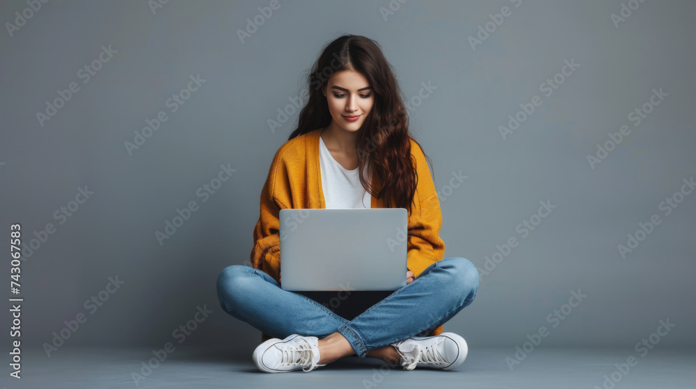 A young woman in a yellow sweater and jeans sits on the floor with her legs crossed, engrossed in her laptop. A young woman in a yellow sweater and jeans sits on the floor with her legs crossed, engrossed in her laptop.