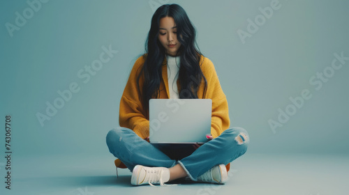 A young woman in a yellow sweater and jeans sits on the floor with her legs crossed, engrossed in her laptop.