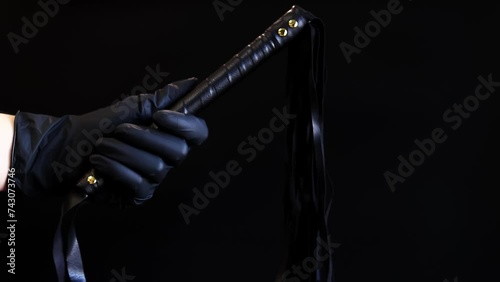 A hand in a black latex glove holds a whip against a dark background. Leather whip for spanking isolated on black. Sex toy for intimacy. Sexual slavery photo
