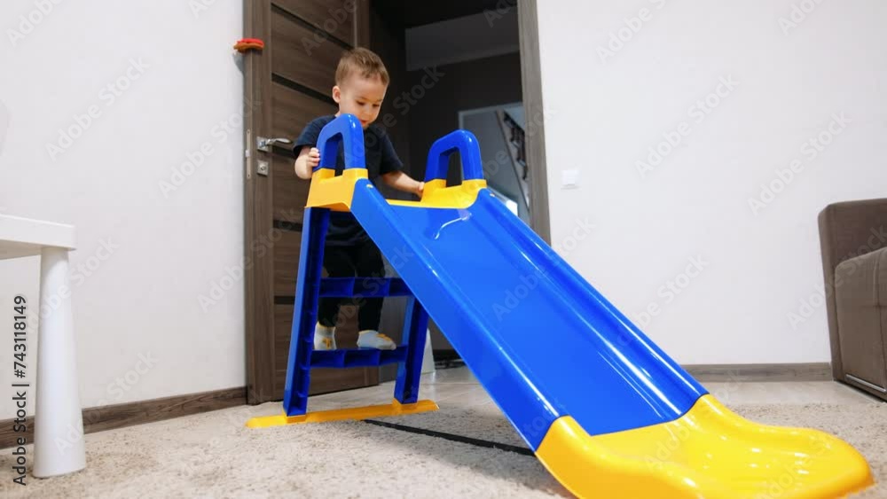 Two year old toddler climbing the slide set in his room. Kid sits on the top for a while and slides down cheerfully.