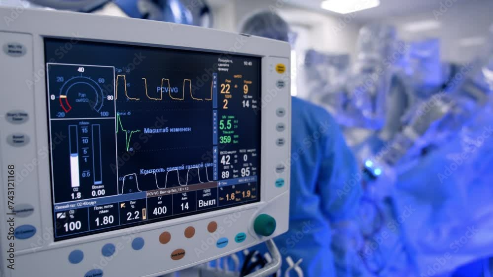 Close look at the screen with life signs of the patient operated in hospital. Robot Da Vinci performs surgery controlled by male surgeon. Blurred backdrop.