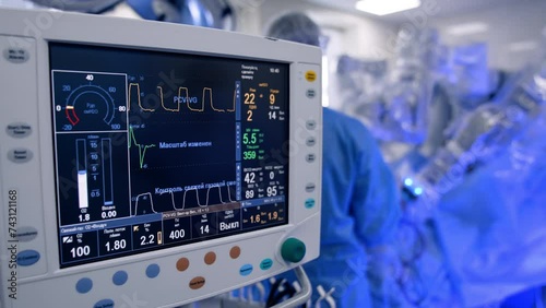 Close look at the screen with life signs of the patient operated in hospital. Robot Da Vinci performs surgery controlled by male surgeon. Blurred backdrop. photo