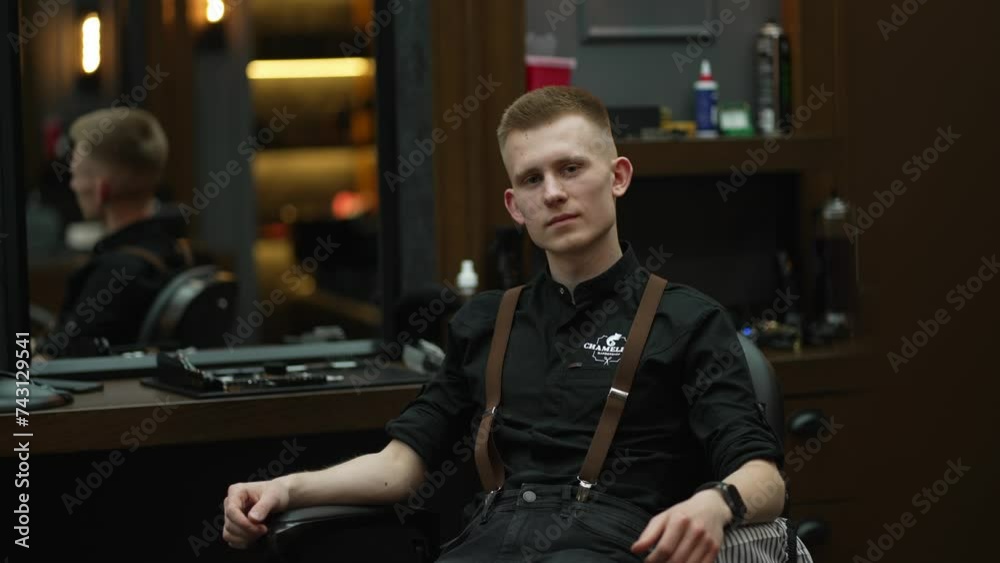 Young hairdresser in professional clothes looks thoughtfully at camera.