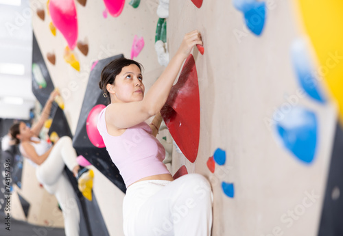 Young Asian woman with difficulty climbs steep artificial wall in sports complex and trains endurance and muscle strength. Challenging obstacle course