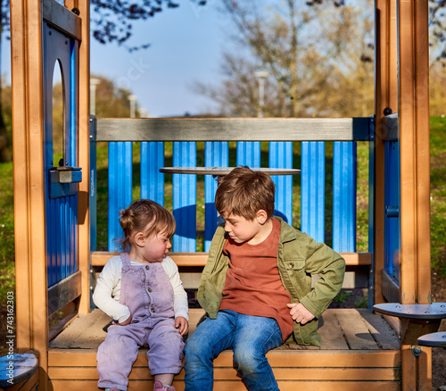 On a sunny day, cheerful children brother and sister looking at each other sitting in a playground. Family love, brotherhood concept with 2 kids siblings looking at one another on a spring day