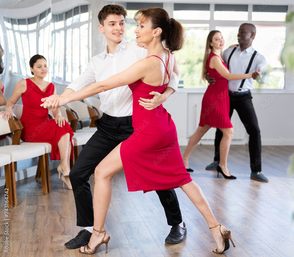 Fototapeta premium Graceful duet, expressive young Hispanic man in formal wear and slender woman in vibrant red dress, engrossed in passionate tango during group dance class in choreographic studio..