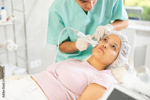 Young male cosmetologist performs hardware facial ultrasound procedure to young female patient