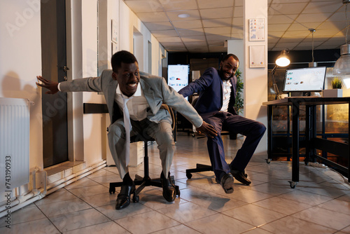 Playful businessmen riding desk chair during sports race competition during break time in modern startup office. Cheerful african american employees having fun  enjoying doing moving activity