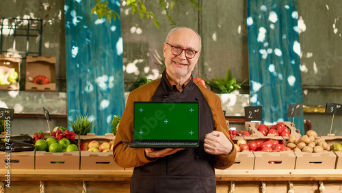 Elderly local farmer holding laptop with isolated greenscreen outdoors at local food fair, sunny day. Senior man showing chroma key display with blank mockup copyspce on screen, wearing pron. photo