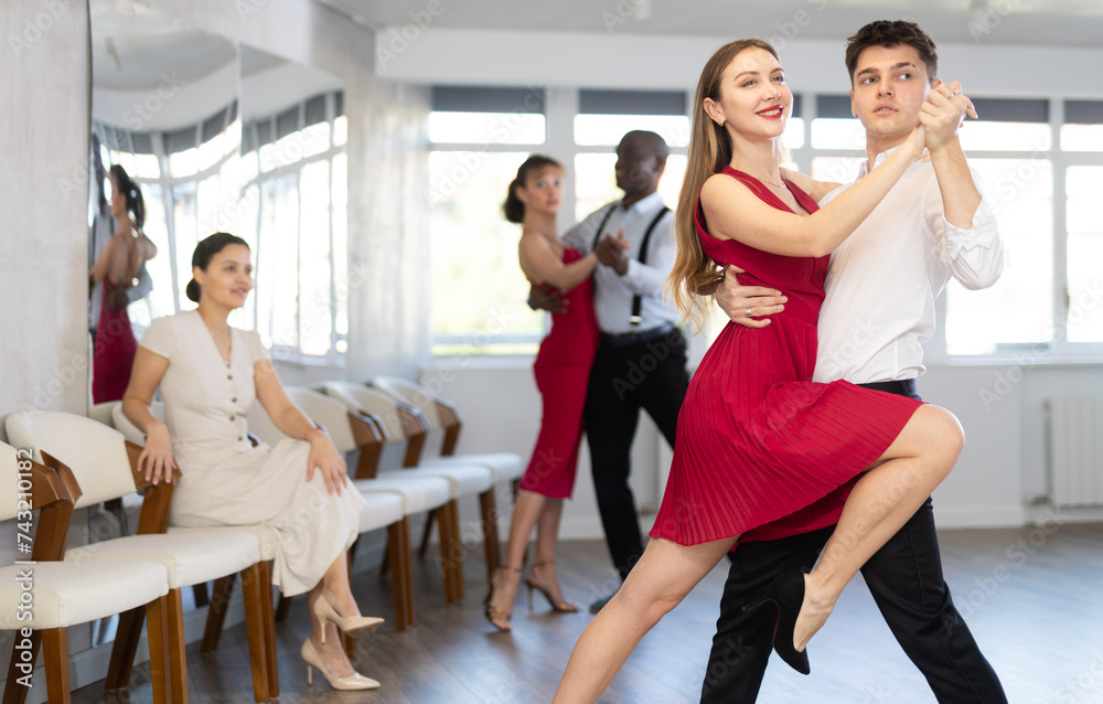 Expressive handsome young guy in formal wear and graceful woman in vibrant red dress rehearsing elegant tango in pair, participating in dance class for adults..