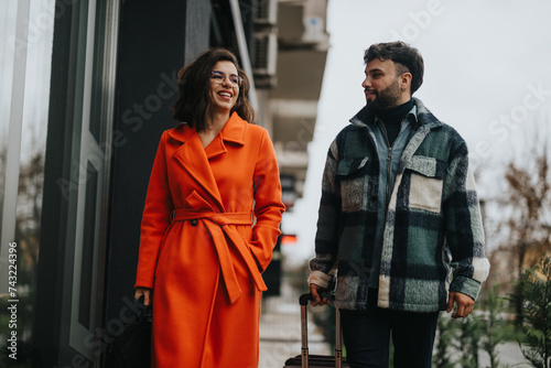 Charming woman in vibrant orange coat laughing with a stylish man during a casual stroll in a city environment, portraying relationship and style.