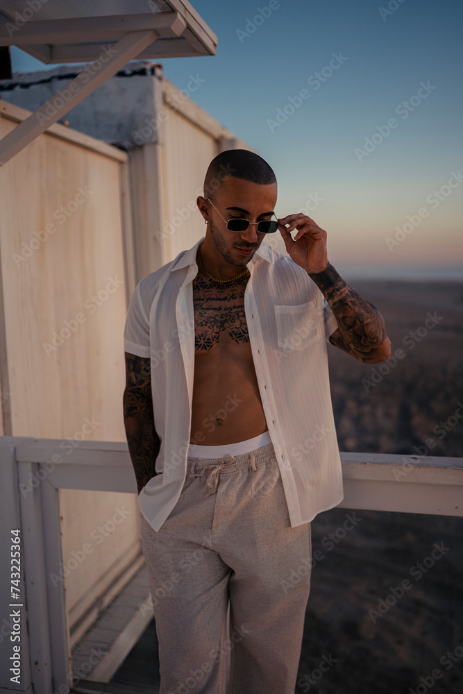 Chico joven tatuado y musculoso posando sin camiseta en la playa al atardecer