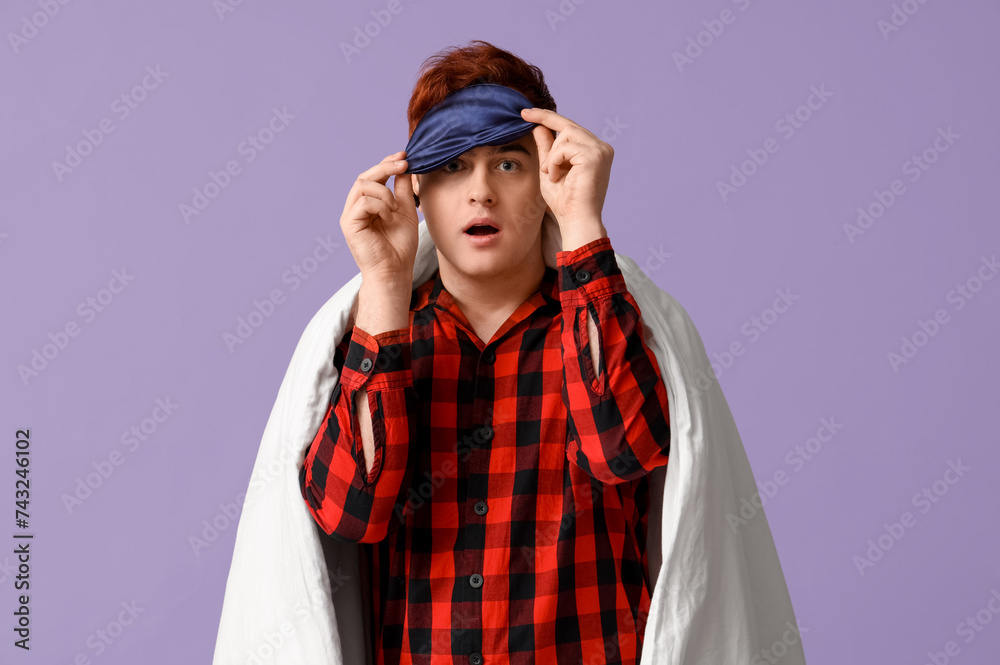 Handsome young shocked man with blanket and sleeping mask on purple background
