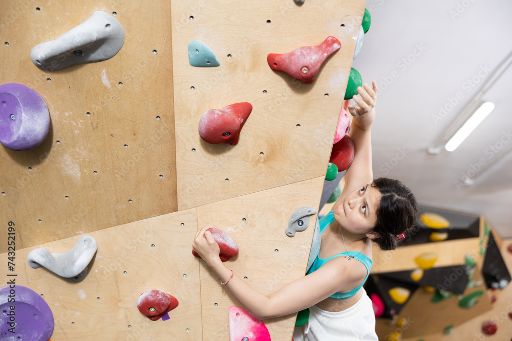 Asian girl is engaged in fitness clambing in modern equipped gym. Girl visiting sports complex climbs steep artificial wall, top view