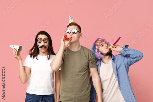 Young friends in funny disguise with party whistles on pink background. April fool's day celebration