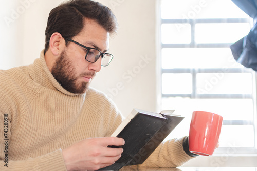 Joven apuesto con estilo tomando café y leyendo un libro en su casa 