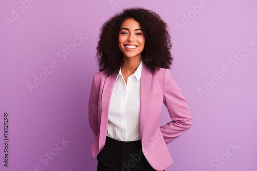 A woman with curly hair and a cheerful smile stands confidently with her hands on her hips in a stylish pink blazer over a white blouse and black trousers, set against a plain purple background