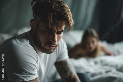 handsome model man sitting on the edge of his bed worried expression, in the background a woman looking at him. relationship problems