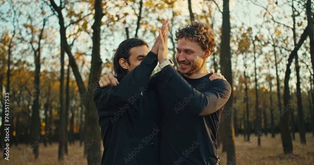 Two happy and cheerful guys posing in the autumn forest after their jog and high-fiving each other. Happy guys after a morning jog look at the camera and pose hugging in black sportswear in the autumn Two happy and cheerful guys posing in the autumn forest after their jog and high-fiving each other. Happy guys after a morning jog look at the camera and pose hugging in black sportswear in the autumn