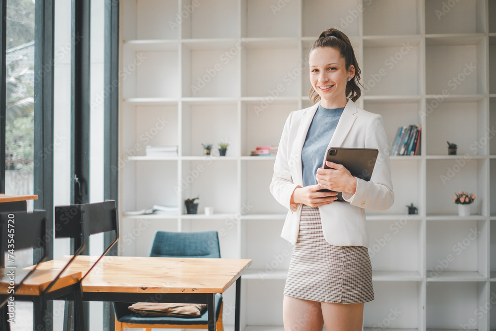 Smiling professional woman holding a tablet stands in a bright, contemporary office space with bookshelves in the background. Smiling professional woman holding a tablet stands in a bright, contemporary office space with bookshelves in the background.