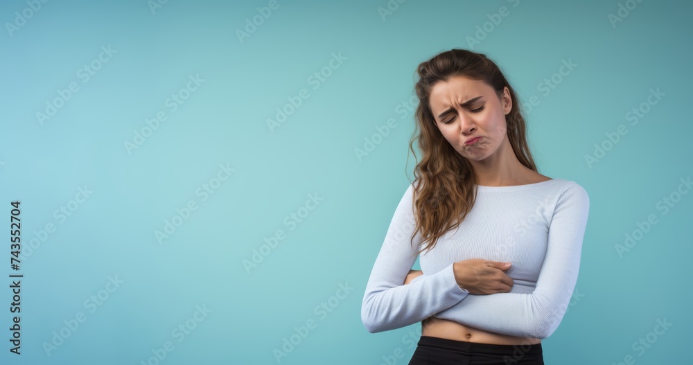 Woman holding her stomach in pain, her face grimacing with discomfort, against a solid light blue background, expressing the distress caused by stomachache - obrazy, fototapety, plakaty 