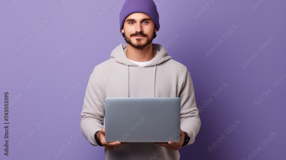 A young successful creative graphic designer, a man wearing a fashionable hoodie and hat, holds a laptop on a purple background with a copy space. A young successful creative graphic designer, a man wearing a fashionable hoodie and hat, holds a laptop on a purple background with a copy space.