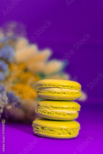 Three bright yellow, appetizing macaroons on a purple background with a bouquet of flowers in the background.