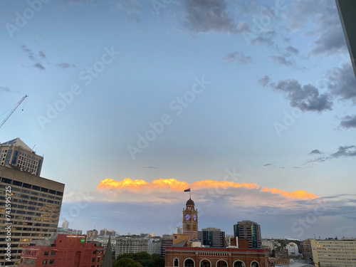 An urban skyline under a vast sky, with clouds illuminated by the warm glow of a setting sun, exuding a calm evening atmosphere.