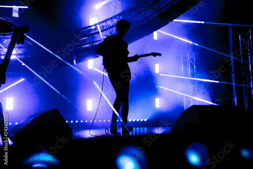 A guitarist captivates the crowd under a vast canvas of blue lasers at an electronic rock concert, delivering a mesmerizing solo performance
