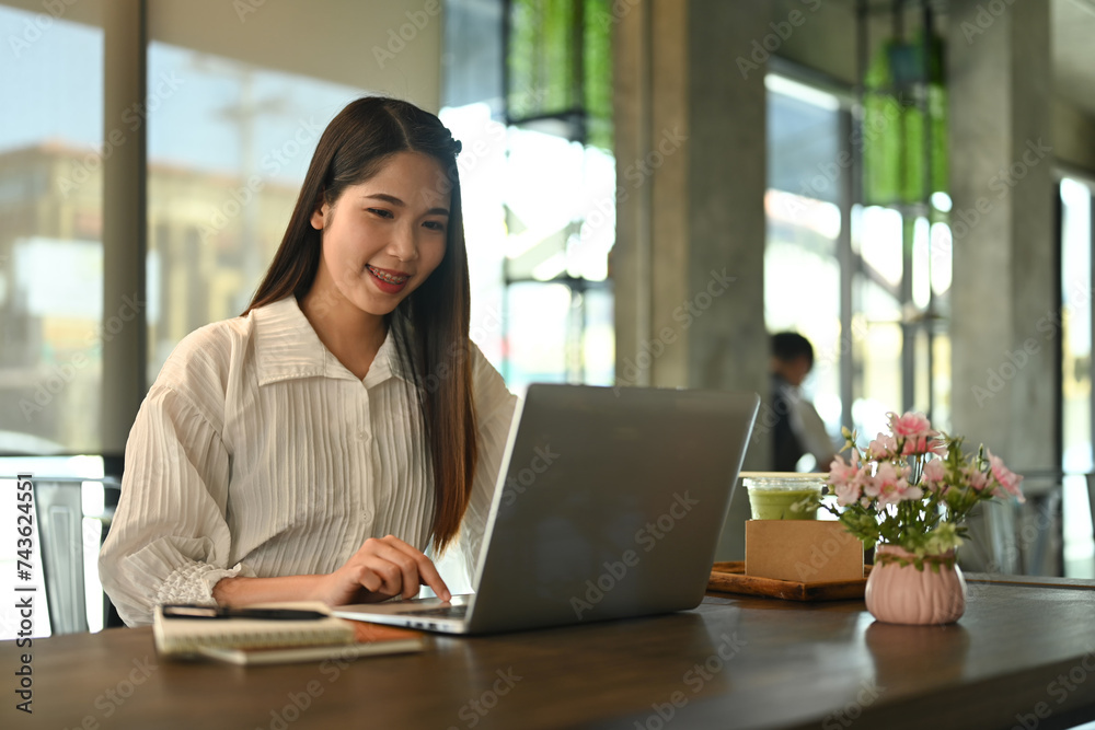 Smiling businesswoman using laptop on wooden table at coffee shop during working remotely Smiling businesswoman using laptop on wooden table at coffee shop during working remotely