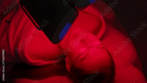 High-angle view of young woman client having red LED light facial photodynamic therapy treatment in modern beauty salon. Luxury medical clinic using phototherapy device with red light wavelength. photo